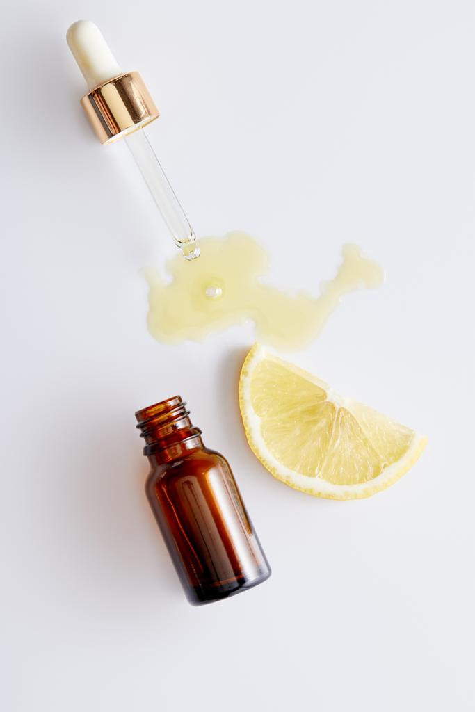 Vue de dessus de l'huile cosmétique qui coule du compte-gouttes à côté de la bouteille et de la tranche de citron sur fond blanc
 - Photo, image