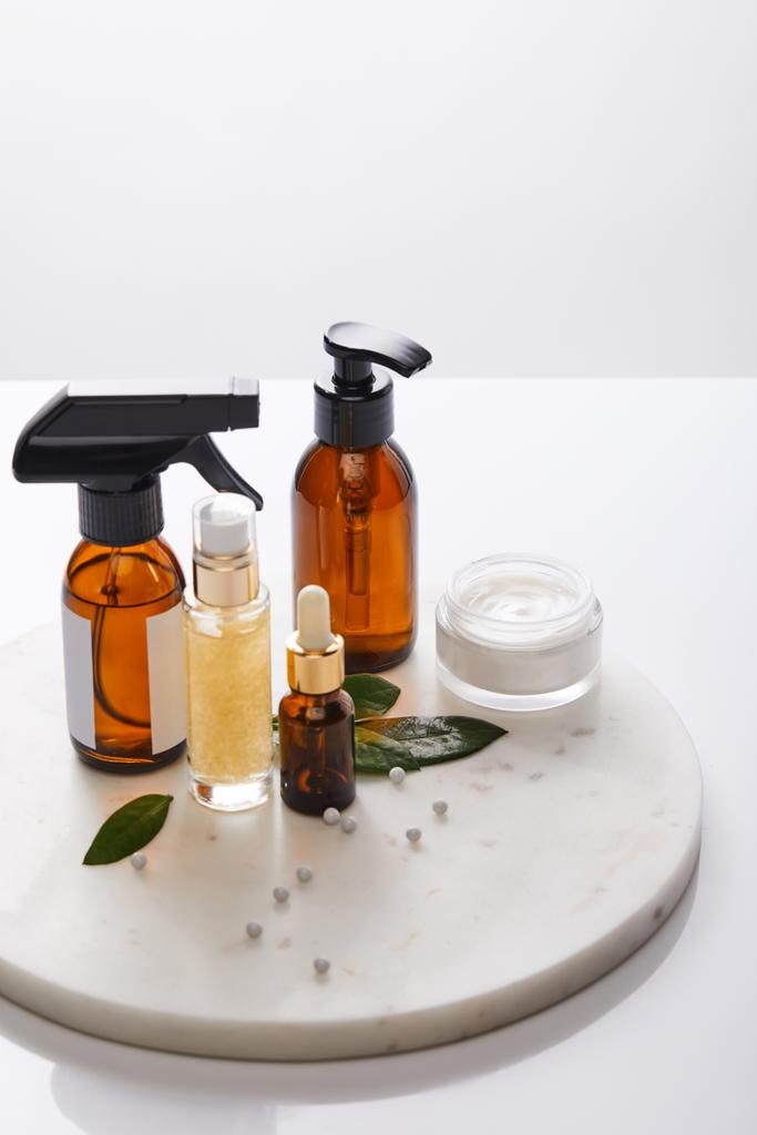 Dispenser bottles of oil and cosmetic cream with leaves and decorative beads on round stand isolated on grey  - Photo, Image
