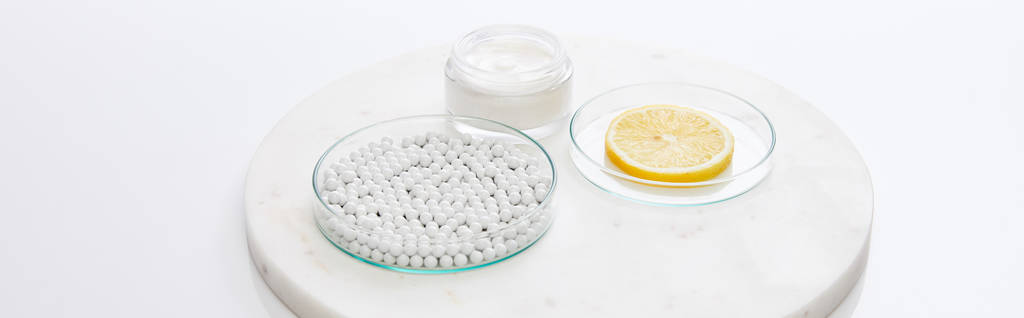 Laboratóriumi üvegáru díszítő gyöngyökkel, citrom szelet kozmetikai krém mellett kerek állványon fehér alapon, panorámás felvétel - Fotó, kép