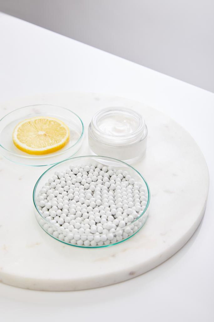 Laborgläser mit Zierperlen und Zitronenscheibe neben Kosmetikcreme auf rundem Ständer isoliert auf grau - Foto, Bild