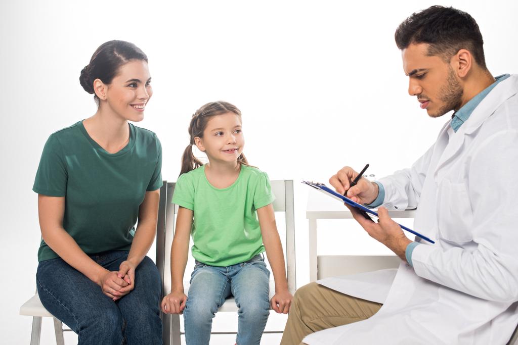 Smiling mother with daughter looking at pediatrician with clipboard isolated on white - Photo, Image