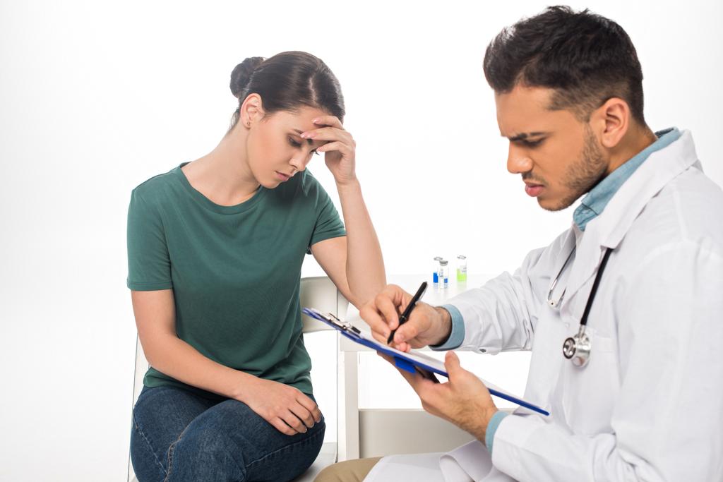 Doctor schrijven op klembord in de buurt trieste patiënt op geïsoleerd op wit - Foto, afbeelding