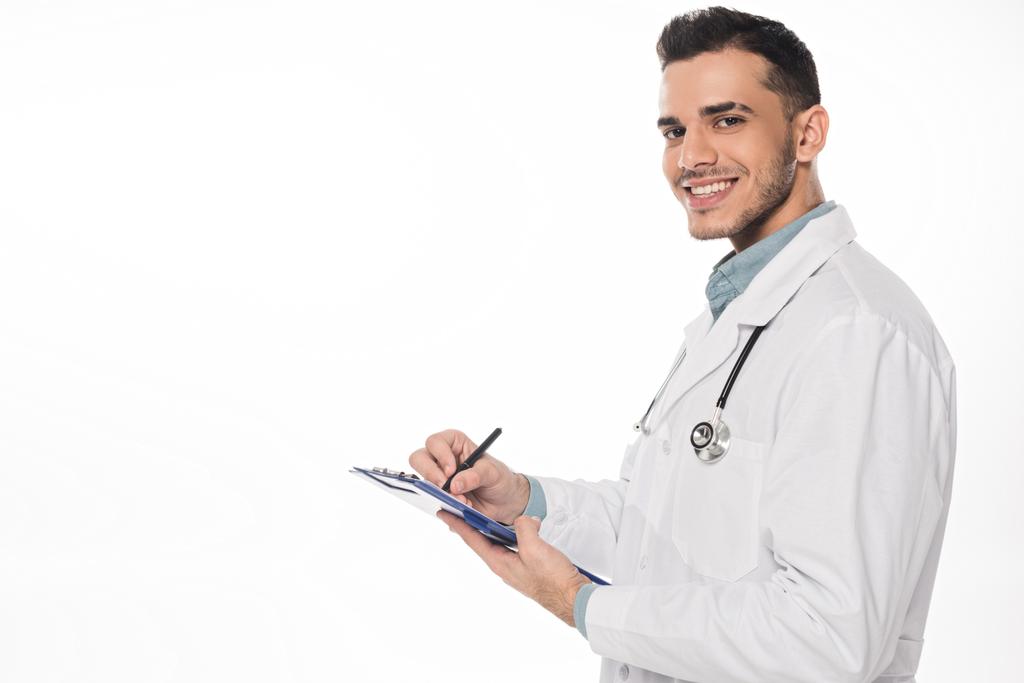 Vista lateral del médico sonriente con portapapeles mirando a la cámara aislada en blanco
 - Foto, imagen