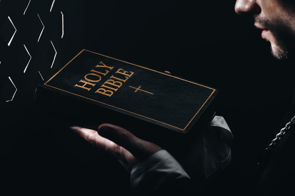 καλλιεργημένη άποψη του καθολικού ιερέα κρατώντας Αγία Γραφή κοντά εξομολογητική πλέγμα στο σκοτάδι με ακτίνες του φωτός - Φωτογραφία, εικόνα