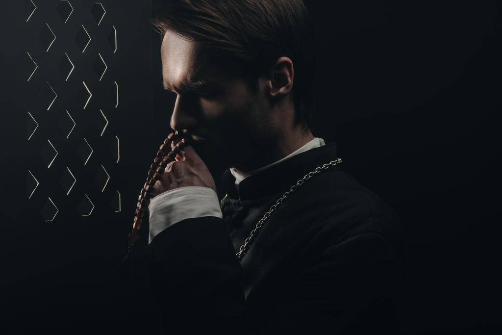 young thoughtful catholic priest kissing wooden rosary beads near confessional grille in dark with rays of light - 写真・画像