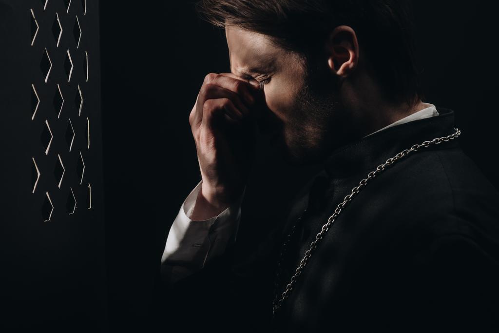 νεαρός τεταμένος καθολικός ιερέας αγγίζοντας το πρόσωπο με κλειστά μάτια κοντά στην εξομολογητική σχάρα στο σκοτάδι με ακτίνες φωτός - Φωτογραφία, εικόνα