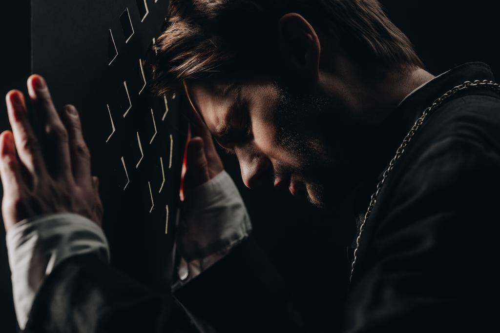 young tense catholic priest with closed eyes leaning on confessional grille in dark with rays of light - Photo, Image