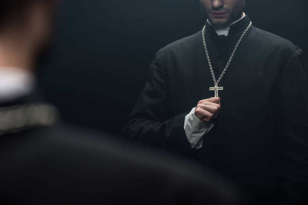 bijgesneden beeld van katholieke priester aanraken kruis op zijn ketting in de buurt van eigen reflectie geïsoleerd op zwart - Foto, afbeelding