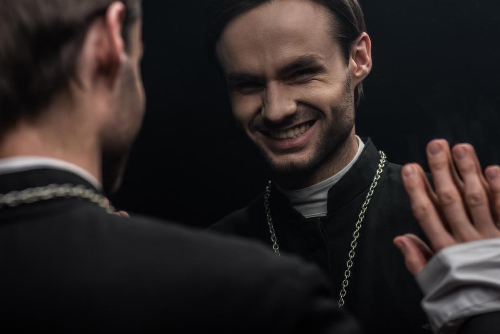 young catholic priest laughing sarcastically while looking at own reflection isolated on black - Fotoğraf, Görsel