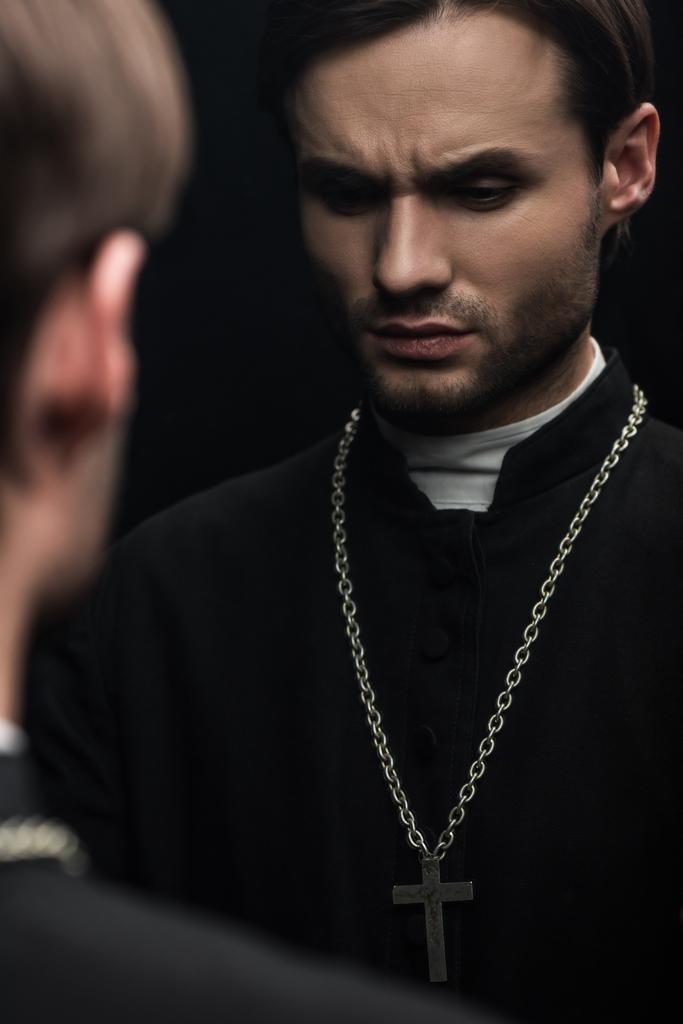 young serious catholic priest thinking while standing near own reflection isolated on black - Fotoğraf, Görsel
