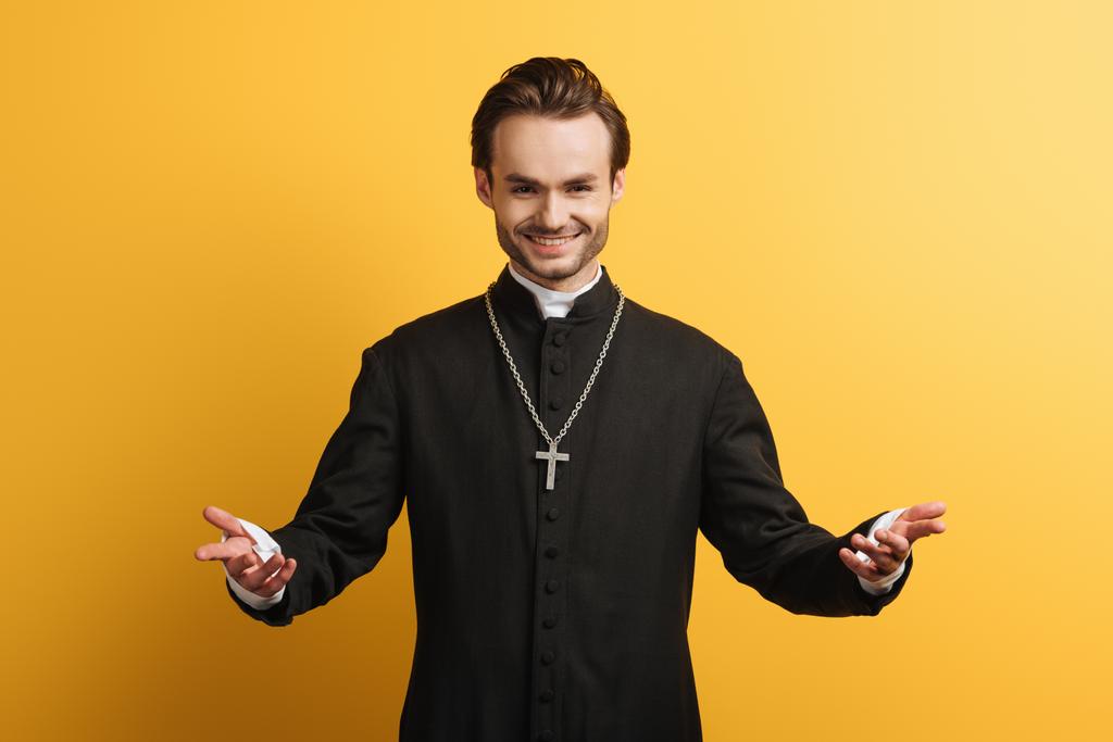 Sacerdote católico sorridente de pé de braços abertos e sorrindo isolado no amarelo
 - Foto, Imagem