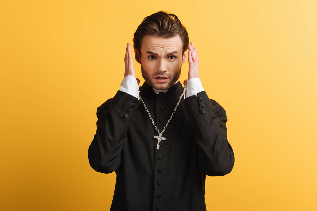 schockierter katholischer Priester hält die Hände in Kopfnähe und blickt isoliert in die Kamera auf gelb - Foto, Bild