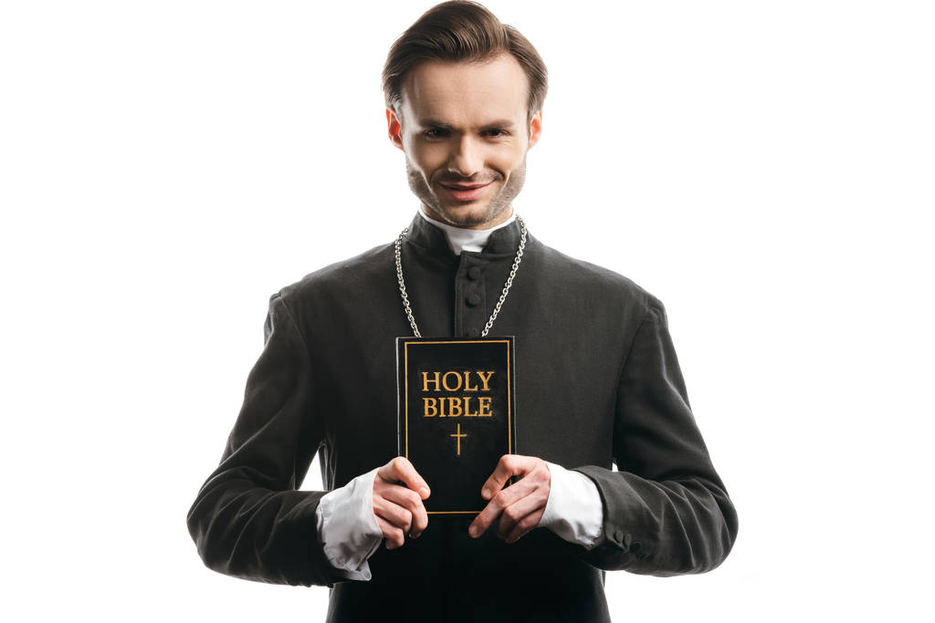 joven sacerdote católico sonriente mostrando la sagrada Biblia aislada en blanco
 - Foto, Imagen