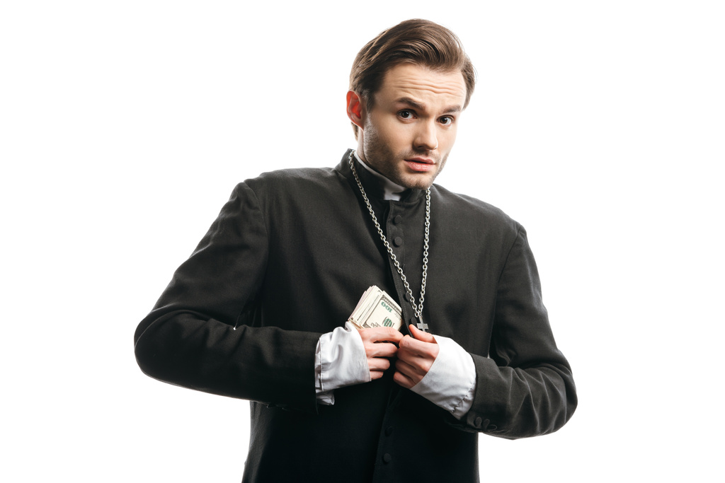 φοβισμένος καθολικός ιερέας κοιτάζοντας κάμερα, ενώ κρύβει χρήματα κάτω από ράσο που απομονώνονται σε λευκό - Φωτογραφία, εικόνα
