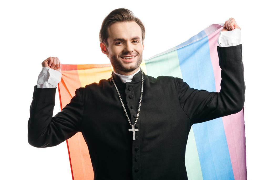 χαμογελαστός καθολικός ιερέας που κοιτάζει την κάμερα ενώ κρατά την σημαία Igbt απομονωμένη στα λευκά - Φωτογραφία, εικόνα