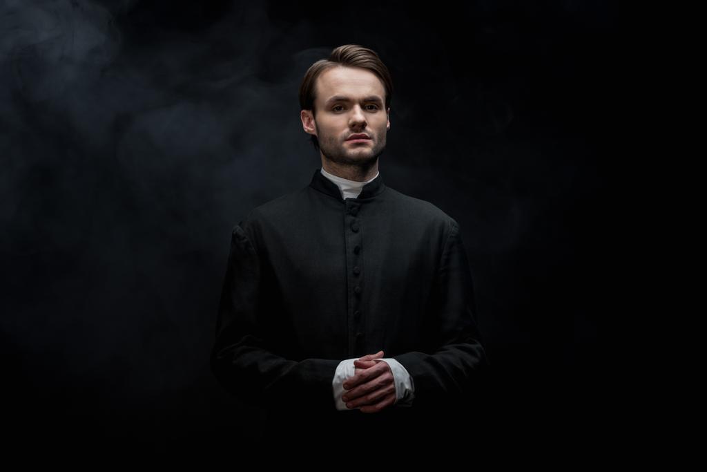 giovane sacerdote cattolico serio in piedi con le mani incrociate su sfondo nero con fumo
 - Foto, immagini