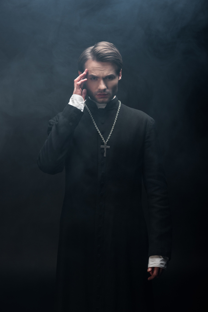 молодий вдумливий католицький священик торкається голови, дивлячись на камеру на чорному тлі з димом
 - Фото, зображення