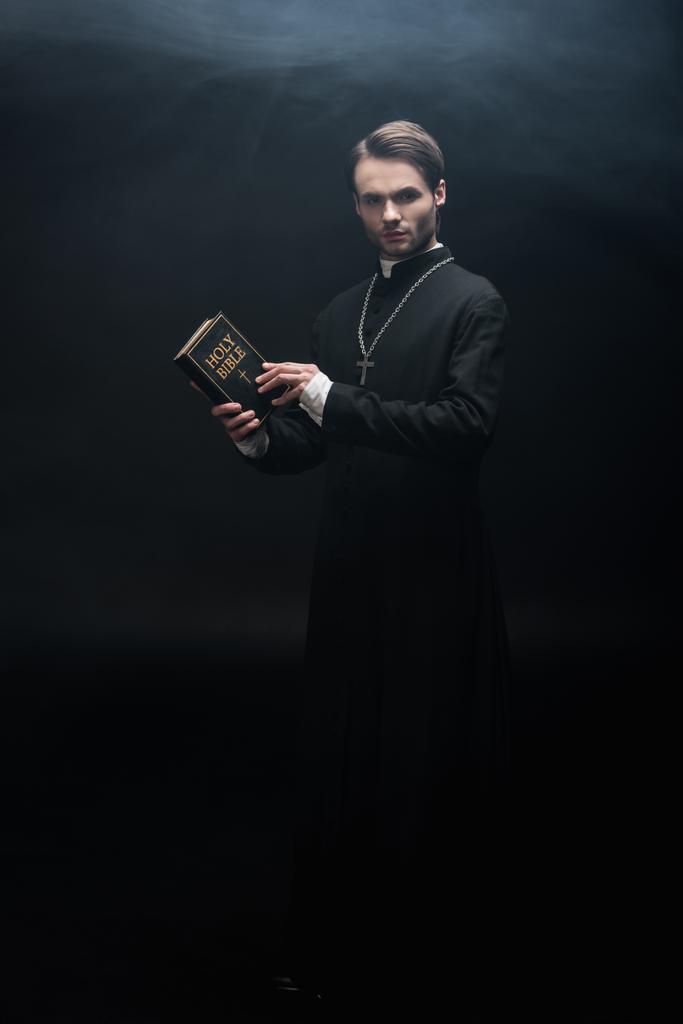 vue pleine longueur du prêtre catholique strict tenant la bible sainte et regardant la caméra sur fond noir avec de la fumée
 - Photo, image