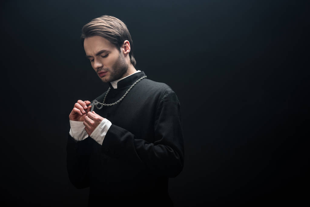 joven sacerdote católico serio mirando la cruz de plata en su collar aislado en negro
 - Foto, Imagen