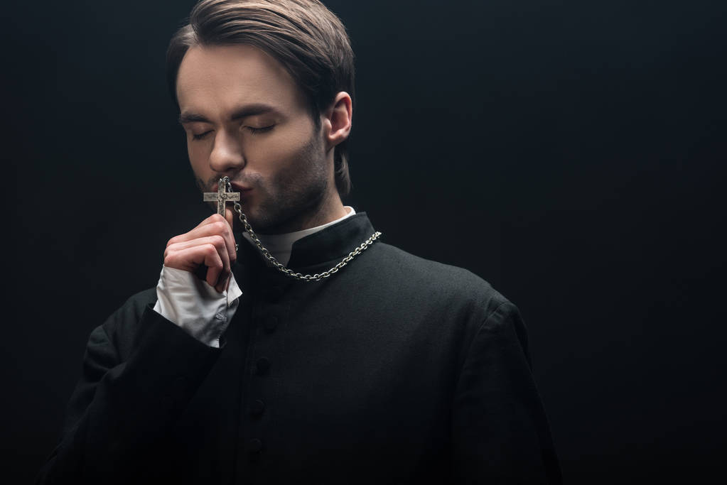 young thoughtful catholic priest kissing silver cross with closed eyes isolated on black - Photo, Image