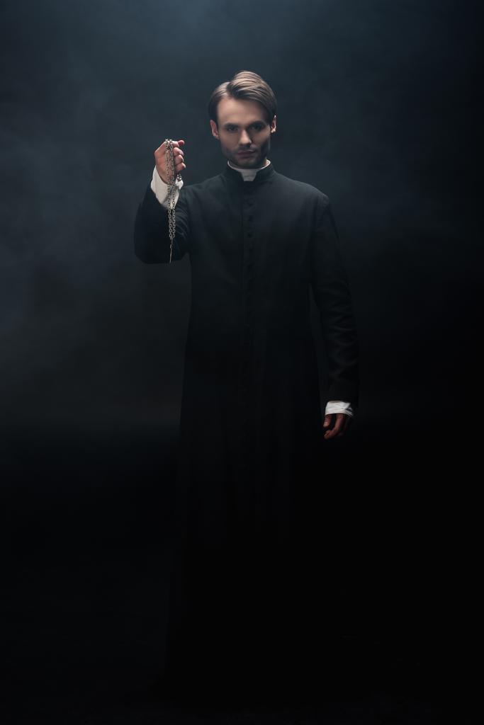 黒を背景に十字架を背景に煙でネックレスを持つ自信に満ちたカトリック聖職者の完全な長距離ビュー - 写真・画像