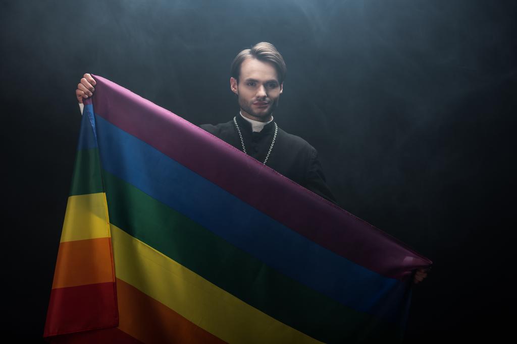 Lächelnder katholischer Priester mit lgbt-Fahne, während er in die Kamera auf schwarzem Hintergrund mit Rauch blickt - Foto, Bild