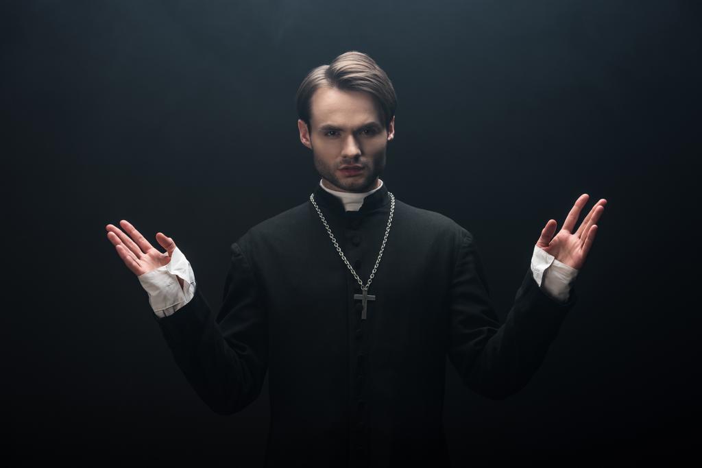 joven sacerdote católico confiado mirando a la cámara mientras está de pie con los brazos abiertos aislados en negro
 - Foto, Imagen