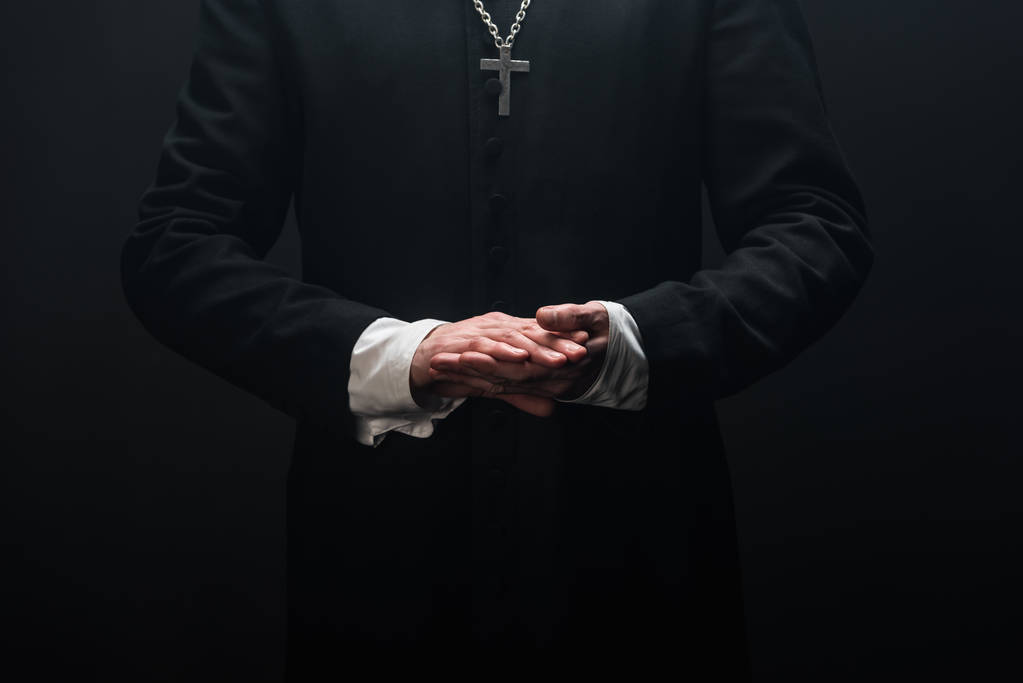 κομμένο από καθολικό ιερέα που στέκεται με διπλωμένα χέρια απομονωμένα σε μαύρο - Φωτογραφία, εικόνα