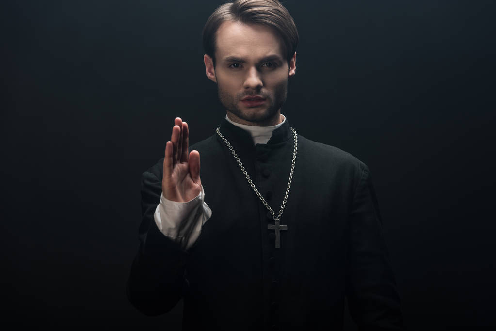 mladý sebevědomý katolický kněz dívá na kameru a ukazuje požehnání gesto izolované na černé - Fotografie, Obrázek