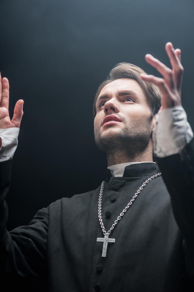 low angle view of young serious catholic priest praying with raised hands isolated on black - Photo, Image