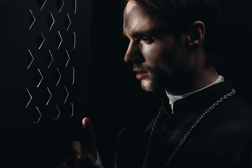 νεαρός τεταμένος καθολικός ιερέας που κοιτάζει μέσα από εξομολογητική γρίλια στο σκοτάδι με ακτίνες φωτός - Φωτογραφία, εικόνα