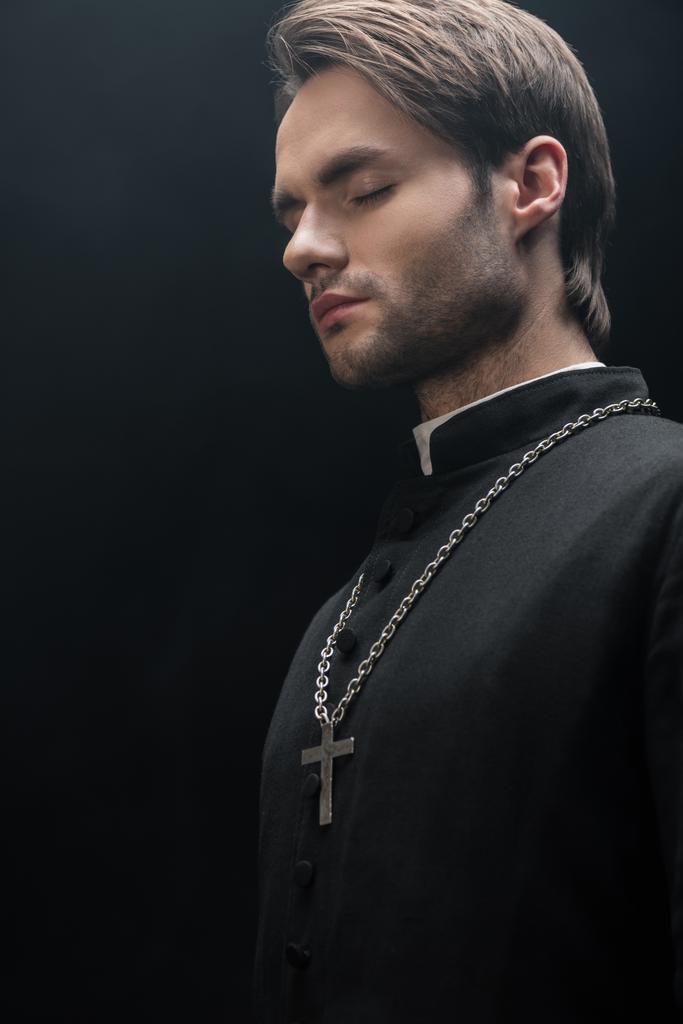 Genç katolik rahip karanlıkta dikiliyor, gözleri kapalı, siyah üzerine izole edilmiş. - Fotoğraf, Görsel