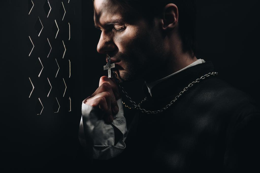 jovem padre católico tenso beijando cruz em seu colar no escuro perto da grade confessional com raios de luz
 - Foto, Imagem