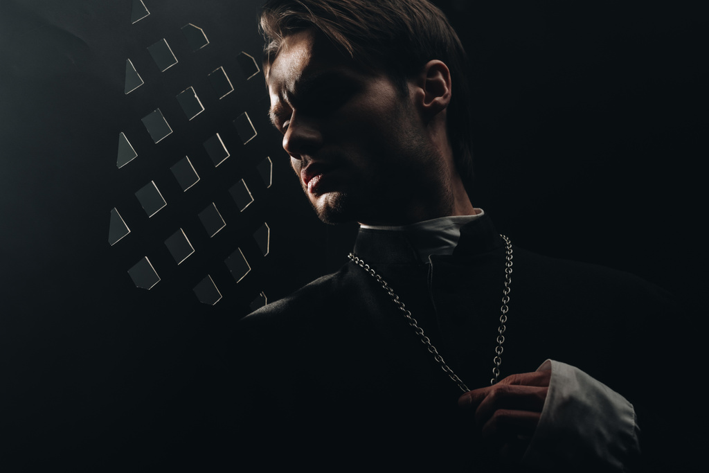 młody poważny katolicki ksiądz dotykający krzyża na naszyjniku w ciemności koło konfesjonału - Zdjęcie, obraz