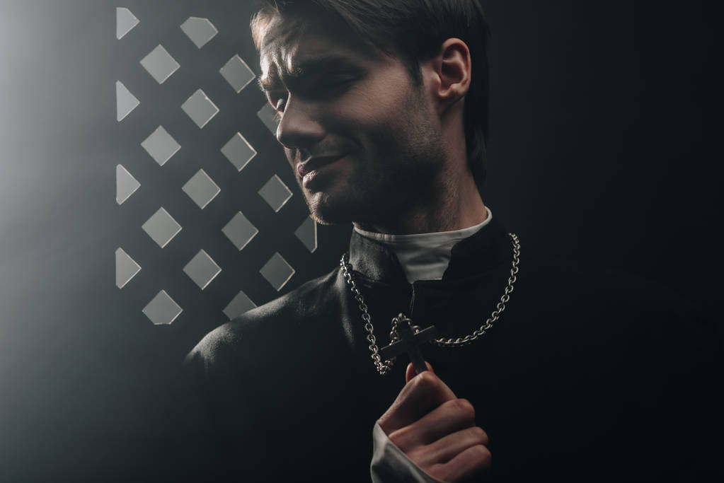jovem padre católico preocupado tocando cruz em seu colar no escuro perto da grade confessional
 - Foto, Imagem