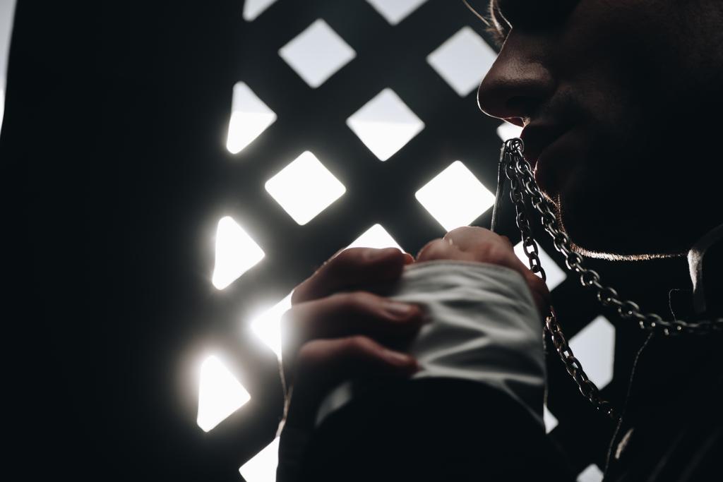 обрезанный вид католического священника целующего крест на своем ожерелье рядом с исповедальней решеткой в темноте с лучами света
 - Фото, изображение