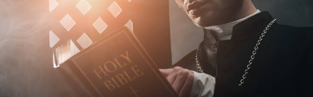 カトリックの聖職者が聖書を読む様子を暗いコンフェディシブルグリルの近くで光とパノラマショットで切り取った - 写真・画像