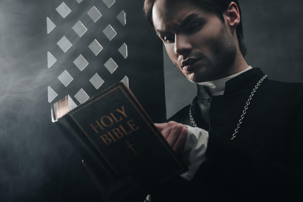 νεαρός στοχαστικός καθολικός ιερέας διαβάζει τη Βίβλο κοντά στο εξομολογητήριο πλέγμα στο σκοτάδι με ακτίνες του φωτός  - Φωτογραφία, εικόνα