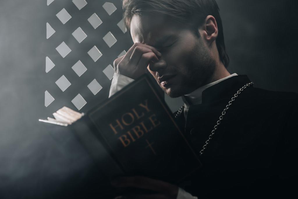 joven sacerdote católico reflexivo tocando la cara mientras lee la biblia cerca de la rejilla confesional en la oscuridad con rayos de luz
 - Foto, Imagen