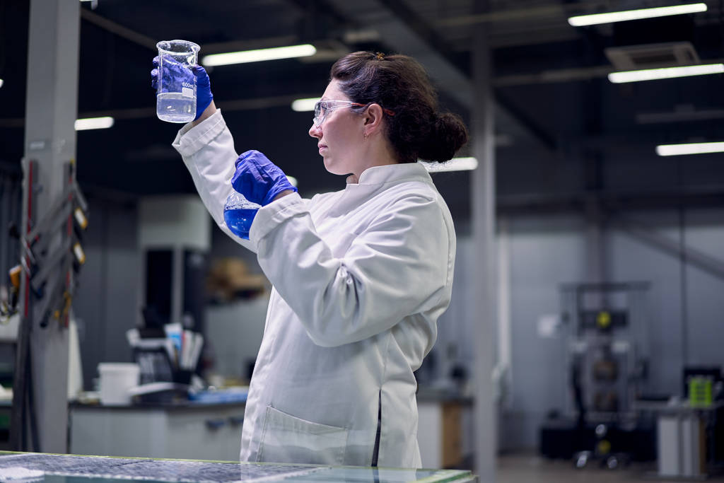 Лаборантка Брюнета в окулярах і білому пальто з блакитною рідиною в руках проводить експерименти - Фото, зображення