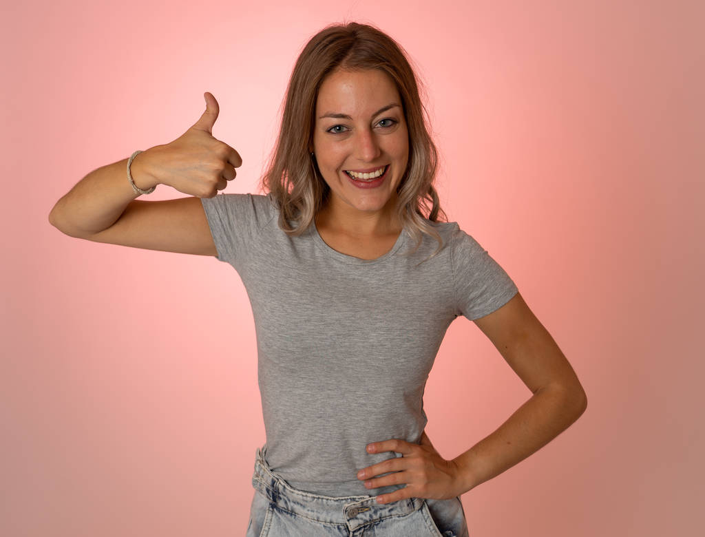 Portrait of beautiful young woman showing thumbs up sign feeling cheerful, happy and satisfied. Young happy student woman making thumb up in joyful approval gesture isolated on pink background. - Photo, Image