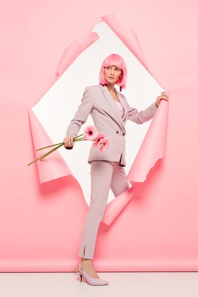 stylish woman in suit and pink wig holding flowers and posing in torn paper, on white - Photo, Image