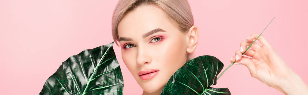 panoramic shot of attractive girl with pink makeup isolated on pink with green leaves  - Photo, Image
