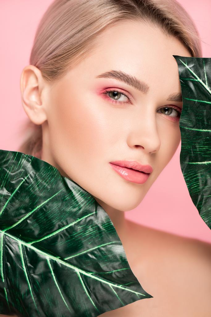 ελκυστική γυναίκα με ροζ μακιγιάζ που απομονώνεται σε ροζ με πράσινα φύλλα  - Φωτογραφία, εικόνα