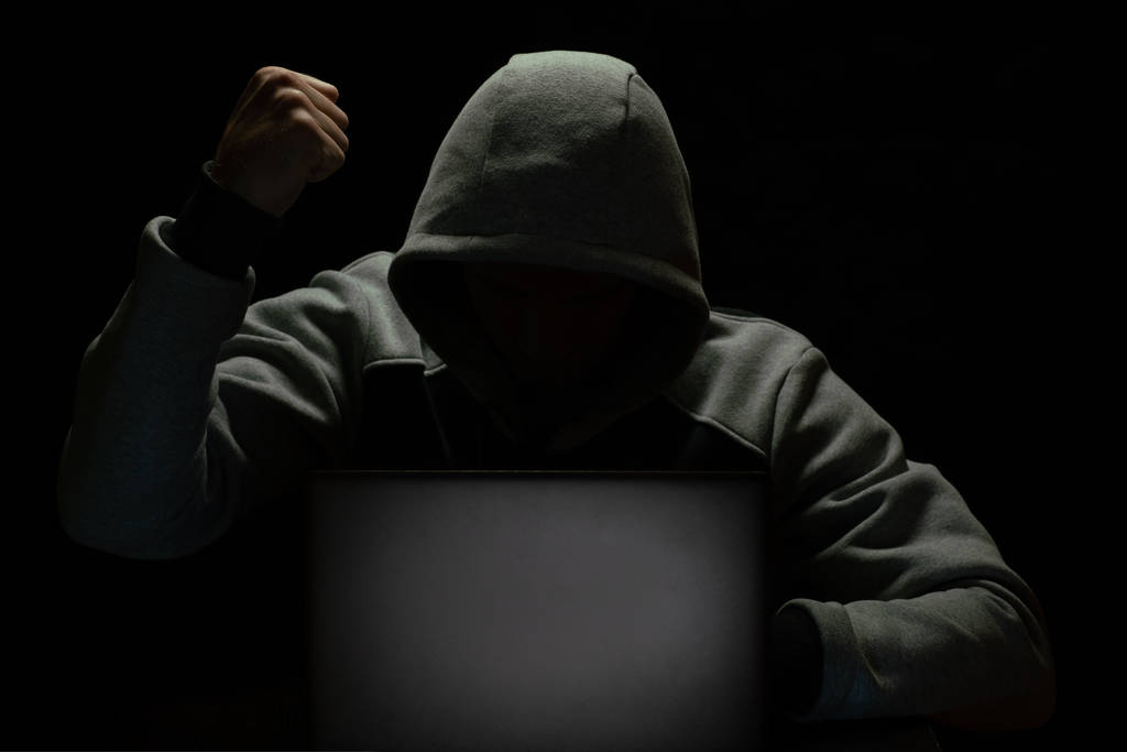 A computer hacker sits in the dark in front of a laptop and is angry, waving his fist - Photo, Image