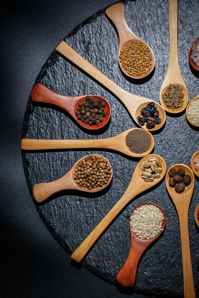 Diferentes especias, pimientos, cilantro, semillas de sésamo y otros para cocinar están en cucharas de madera sobre un fondo negro
 - Foto, Imagen