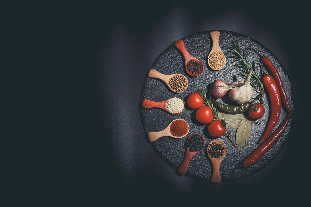 Μπαχαρικά σε ξύλινα κουτάλια και ντοματίνια, πιπεριές τσίλι, σκόρδο και μυρωδικά για μαγείρεμα σε μαύρη επιφάνεια και μαύρο φόντο - Φωτογραφία, εικόνα
