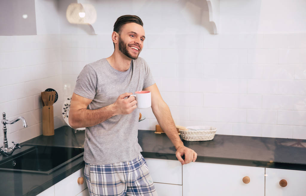 Gelukkig bebaarde man met beker met koffie in de keuken - Foto, afbeelding