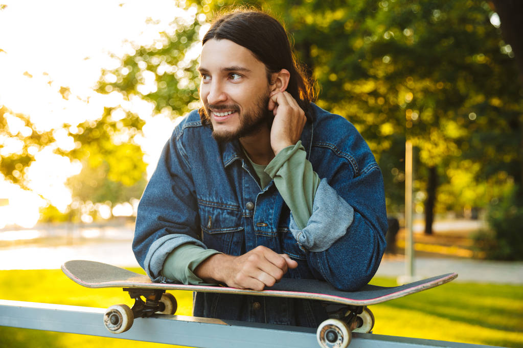 Usmívající se optimistický mladý vousatý muž se skateboardem stojící v přírodním parku. - Fotografie, Obrázek
