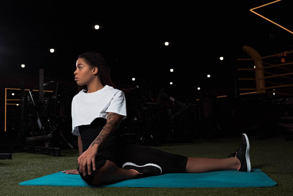 Attractive african american woman sitting and stretching on fitness mat in gym - Photo, Image